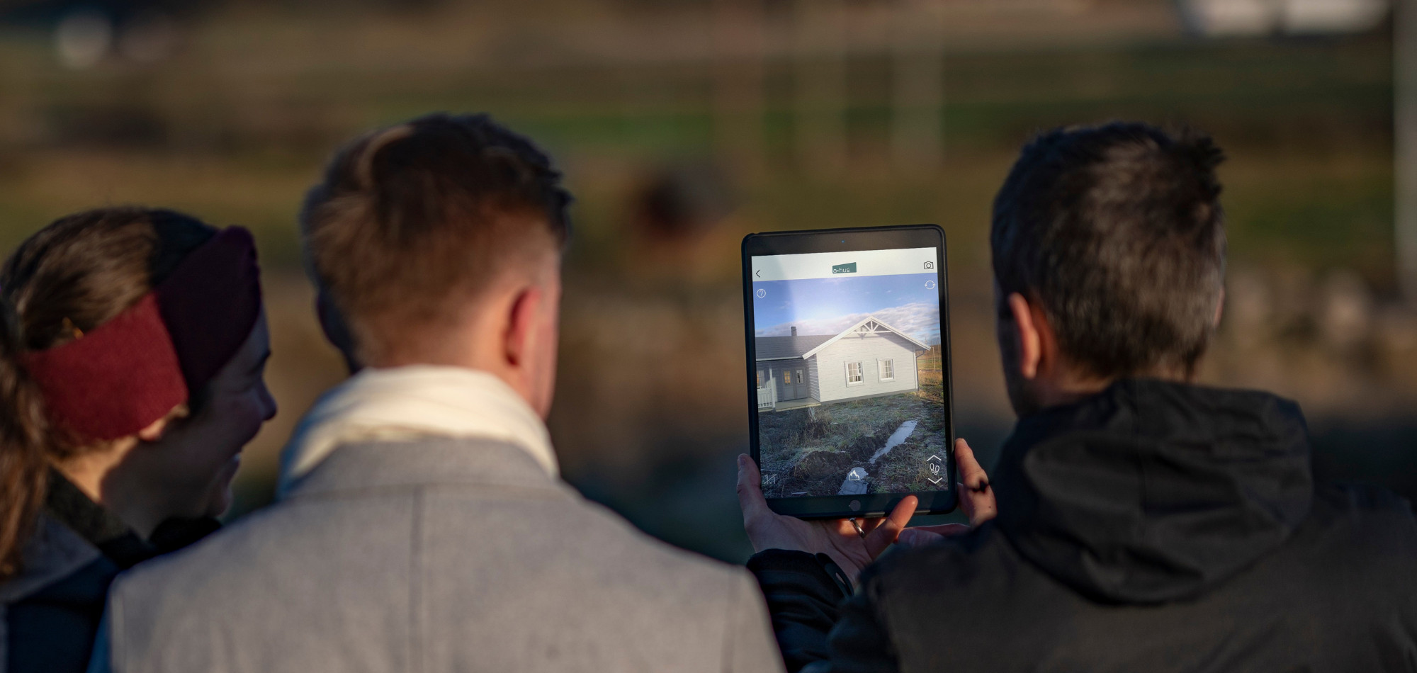 Tre personer tittar på en surfplatta som visar ett virtuellt hus i ett landskap, en illustration av Deromes arbete med trähus och modern teknik.