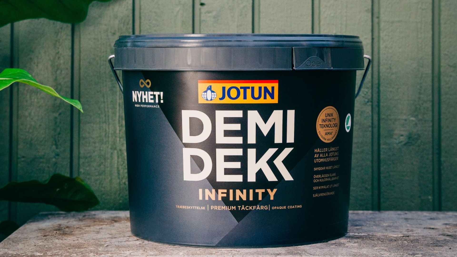  En hink med Jotun Demidekk Infinity täckfärg placerad utomhus framför en grönmålad trävägg.