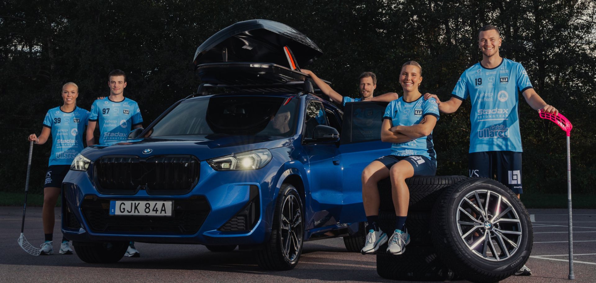 En kampanjbild för Lunds Bils "Winter Edition" där fem innebandyspelare från Warberg IC poserar i blå matchställ tillsammans med en blå BMW utrustad med takbox. Spelarna står i en halvcirkel runt bilen på en parkeringsplats, och i förgrunden syns däckstaplar och innebandyklubbor som knyter an till samarbetet mellan bilhandlaren och idrottsklubben.