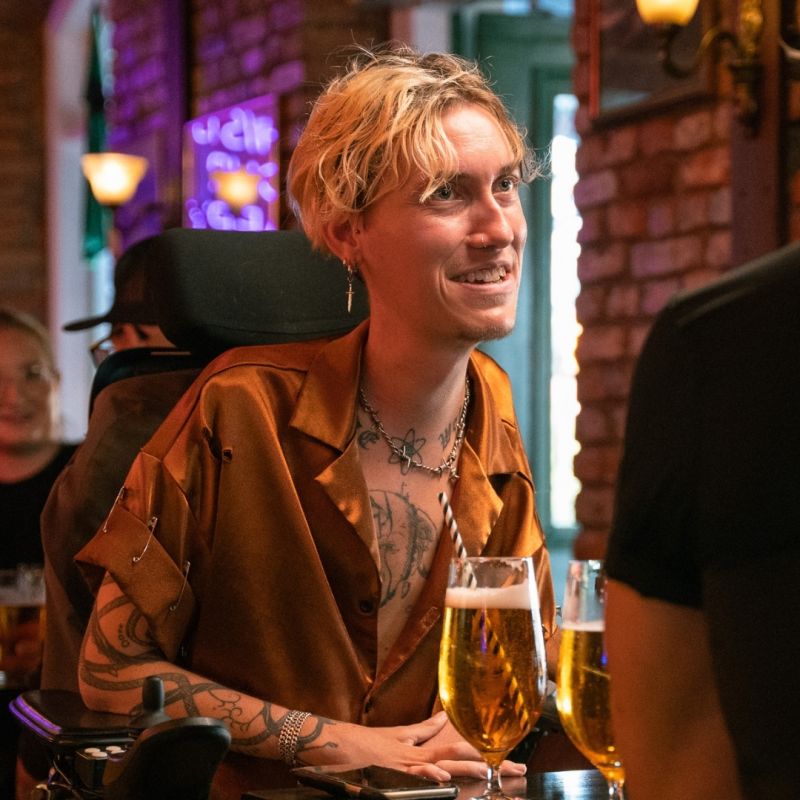  En ung man i rullstol ler varmt under en utekväll på en restaurang eller bar, fångad i ett ögonblick av gemenskap med vänner.