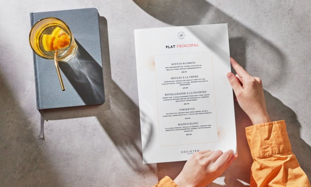  Societéns tryckta meny med den nya grafiska profilen, presenterad tillsammans med en drink i en ljus och modern miljö.