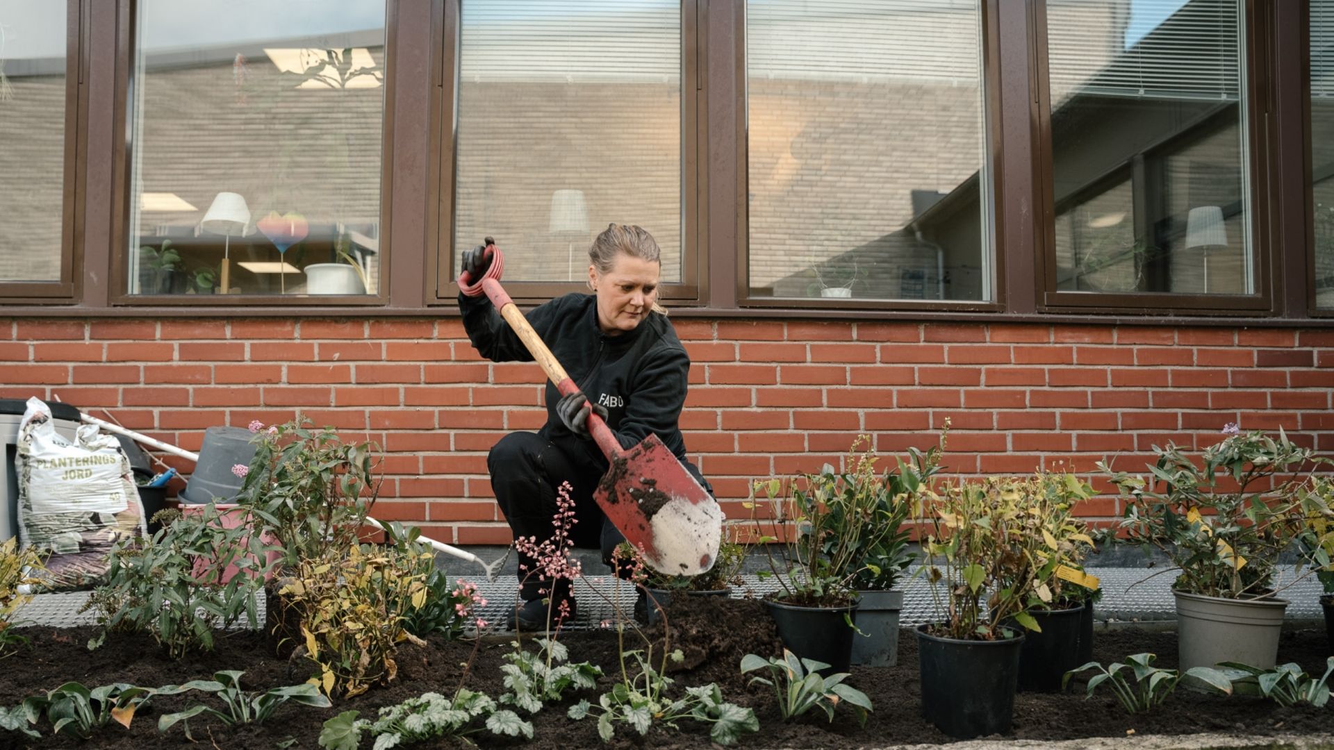  En medarbetare från Fabo använder en spade för att plantera växter i en rabatt utanför en tegelbyggnad, vilket speglar arbetet med att skapa trivsamma utemiljöer.