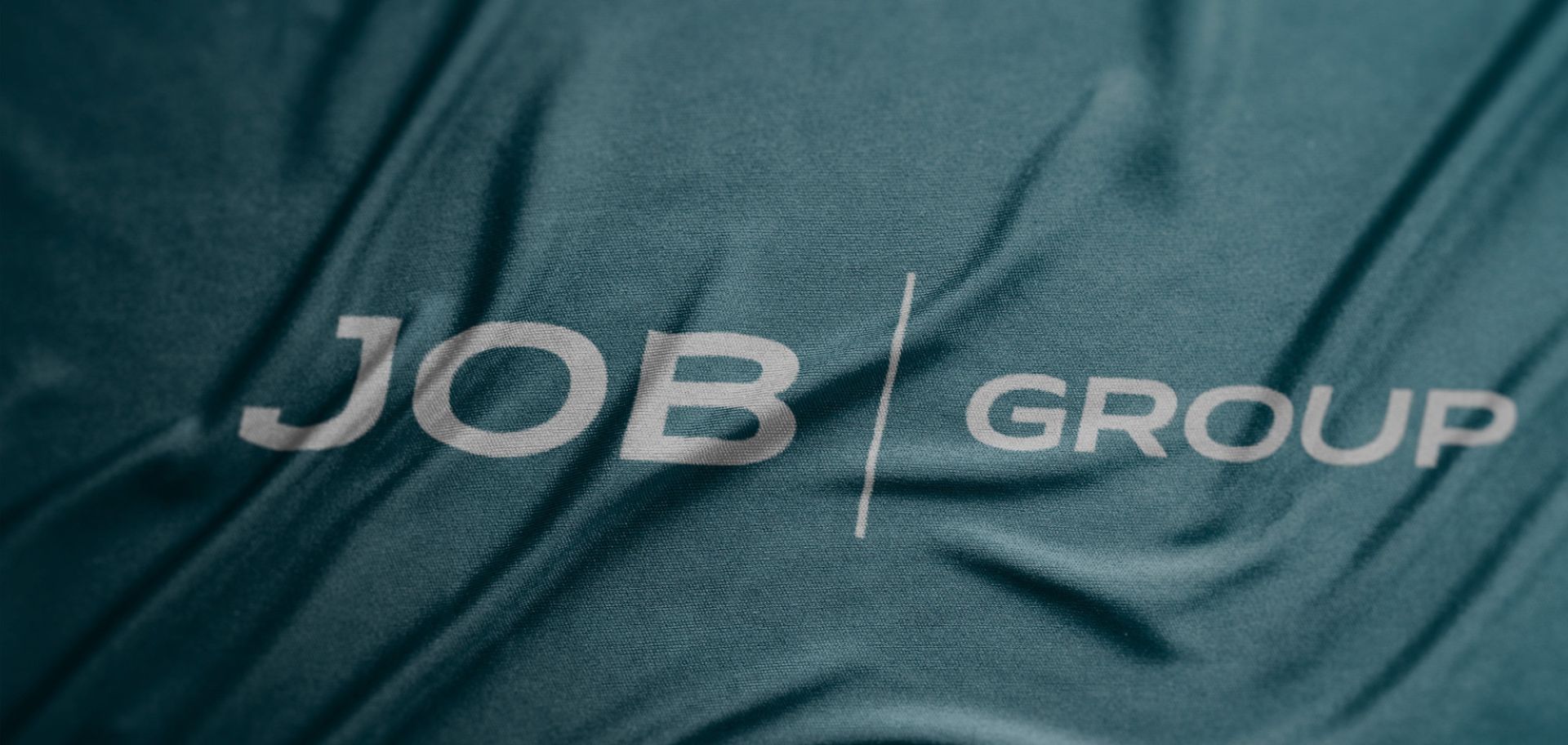 En närbild på JobGroups logotyp tryckt i silvergrått mot ett vågigt, petrolblått tyg.