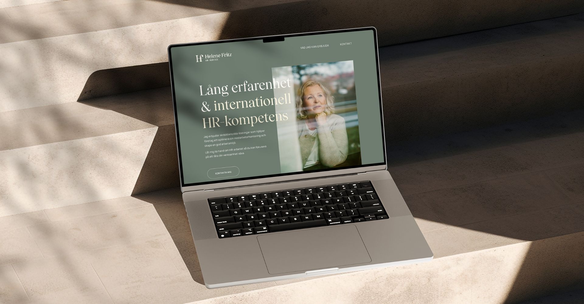 En bärbar dator som visar hemsidan för Helene Fritz HR-Service mot en trappa i betong. Skärmen visar rubriken "Lång erfarenhet & internationell HR-kompetens" och en bild på Helene Fritz mot en dämpad grön bakgrund, badad i naturligt solljus.