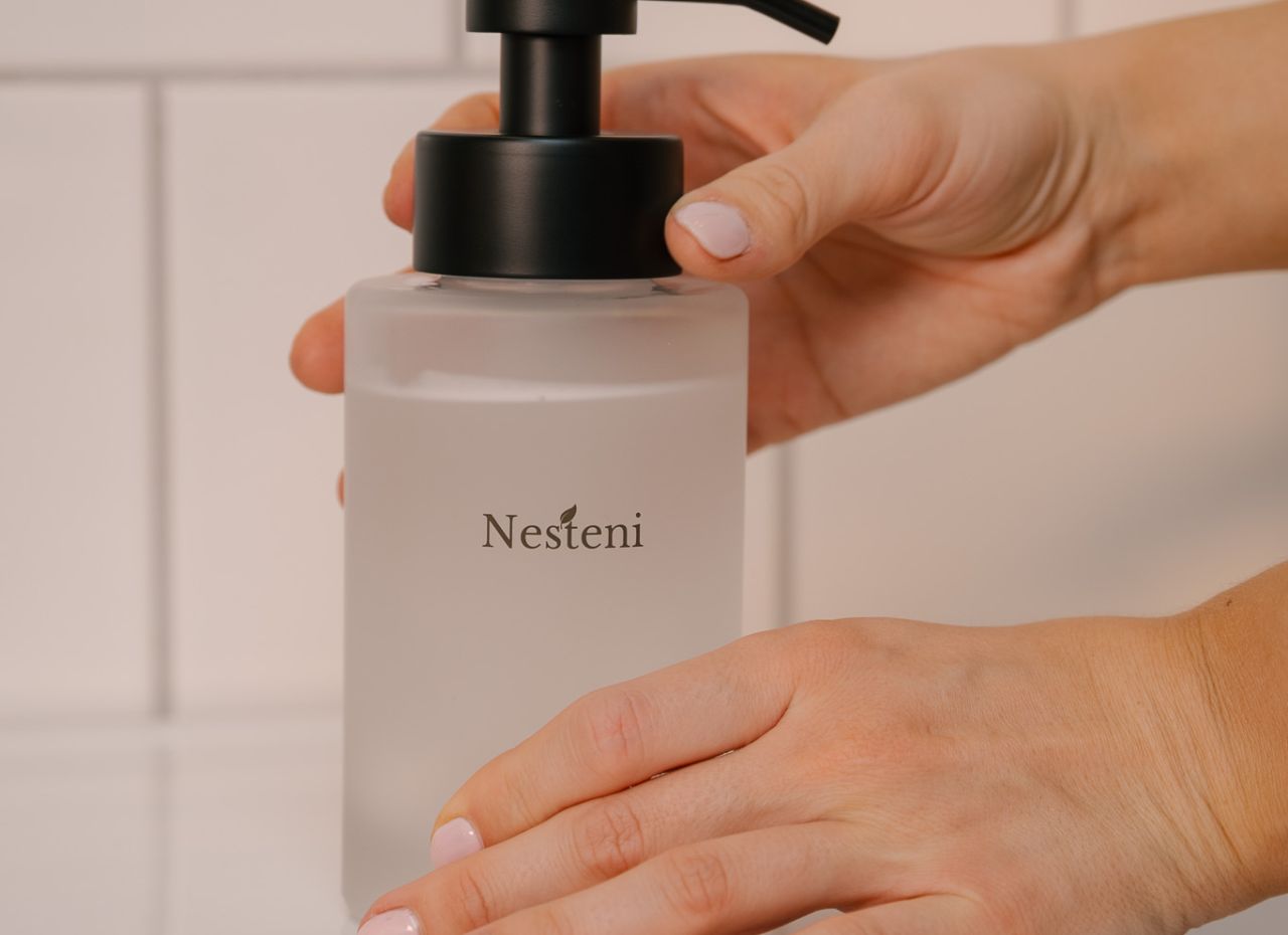 Händer som håller i en tvålpump från Nesteni i frostat glas.