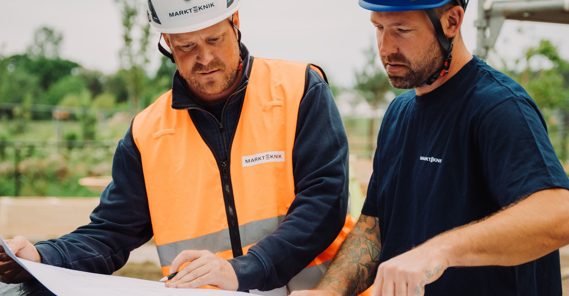  Två medarbetare från Markteknik i arbetskläder studerar en ritning tillsammans på en byggarbetsplats.