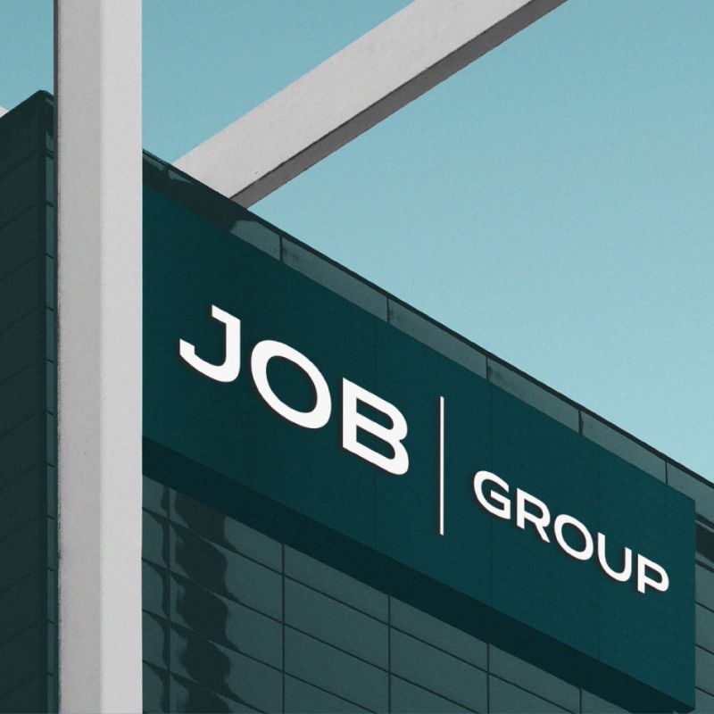 En fasadskylt på en modern byggnad som visar JobGroups logotyp i vitt mot en mörk petrolblå bakgrund under en klarblå himmel.