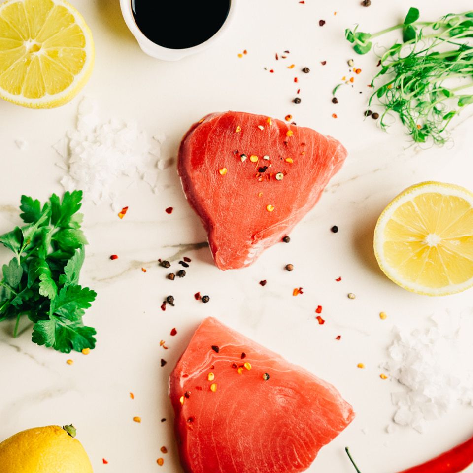  En närbild på två färska tonfiskfiléer på en marmorskiva, omgivna av citroner, flingsalt, örter och kryddor för att framhäva råvarans kvalitet.