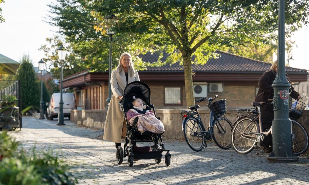  En kvinna går med ett barn i en specialanpassad vagn längs en kullerstensgata i en solig stadsmiljö, en bild som fångar vardagen för familjer i projektet "Toaletter för alla".
