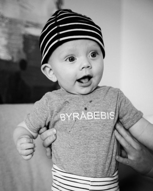 Svartvitt foto av en glad bebis med randig mössa och en tröja med texten 'BYRÅBEBIS'