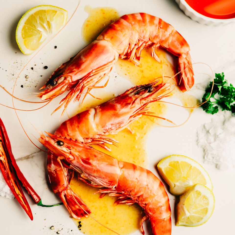 En närbild på tre färska jätteräkor på en marmorskiva, omgivna av citroner, chili och örter för att lyfta fram råvarans kvalitet