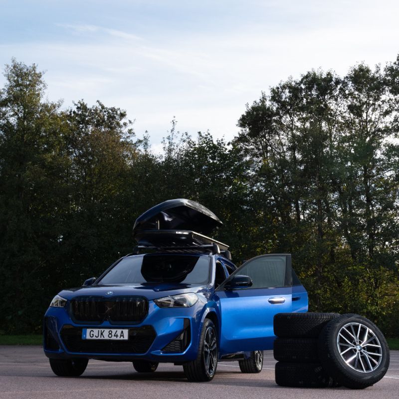 En blå BMW med öppen förardörr och en monterad takbox står parkerad på en asfalterad yta. I förgrunden syns en stapel med vinterdäck, och i bakgrunden ramar en tät vägg av gröna träd in motivet under en ljusblå himmel.