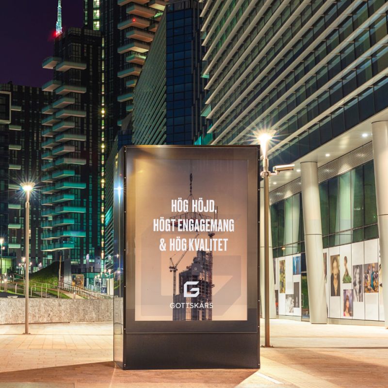 En belyst reklampelare i en modern stadsmiljö kvällstid som visar Gottskärs budskap: ”Hög höjd, högt engagemang & hög kvalitet”. Bakgrunden består av upplysta skyskrapor som speglar företagets expertis inom stora byggprojekt.