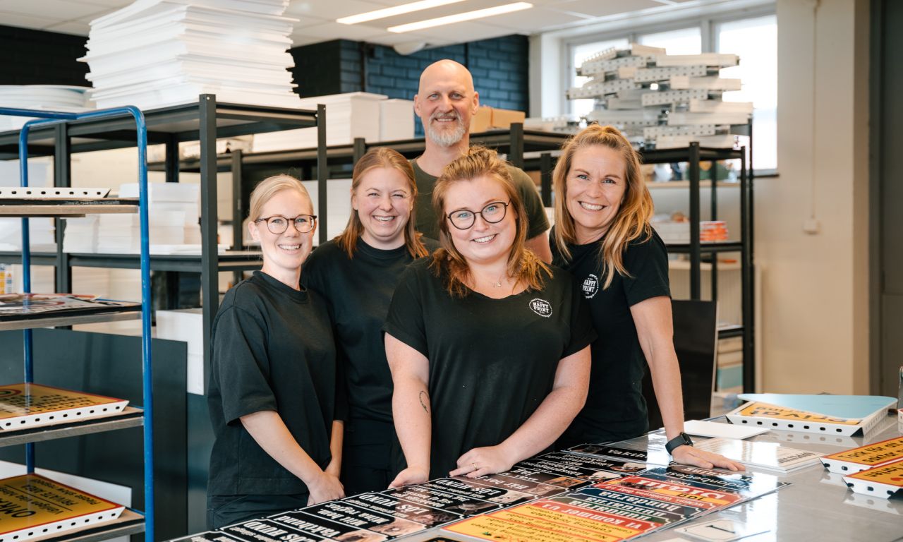 Personal från Happy Print vid ett arbetsbord fyllt med olika varningsskyltar och emaljliknande skyltar.