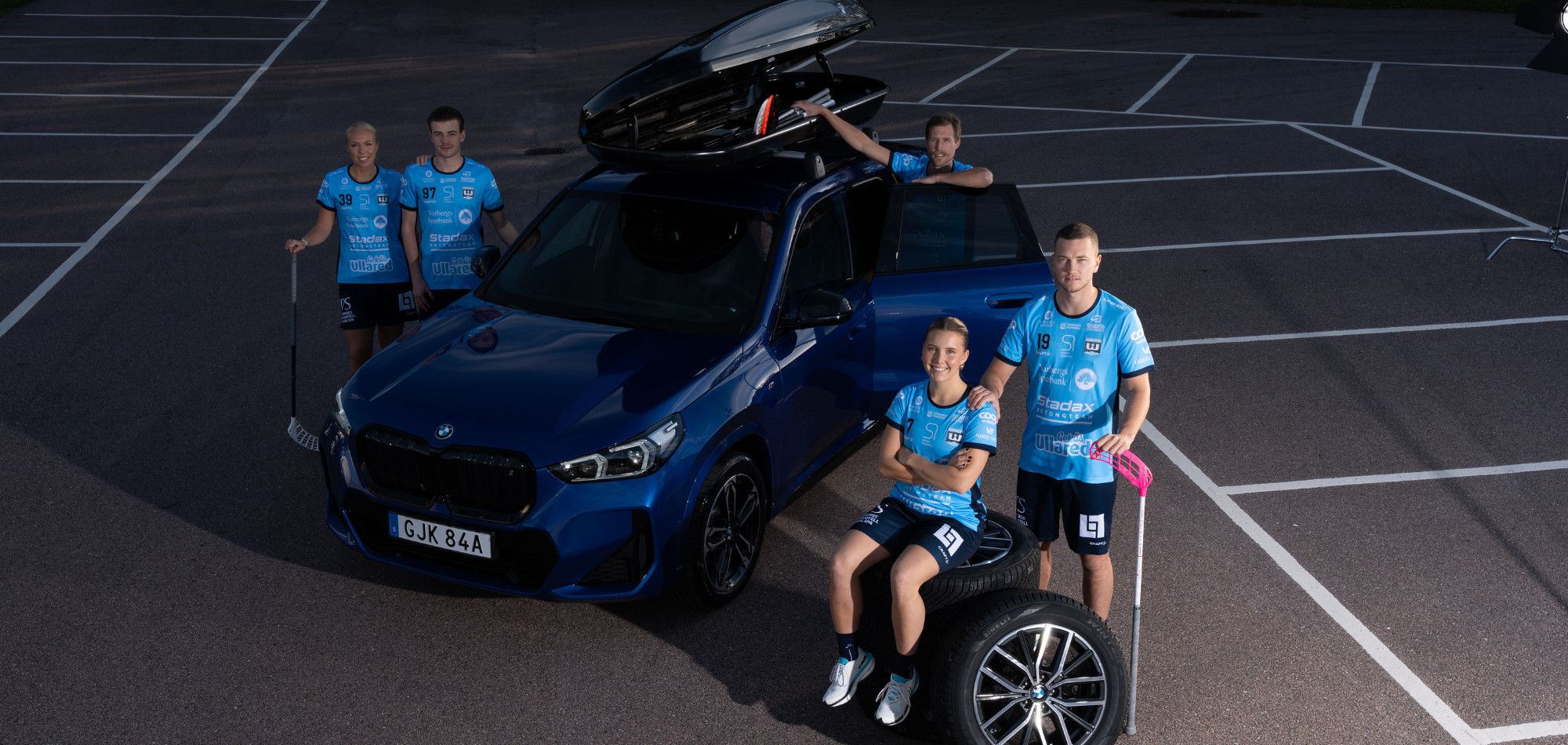 En vidvinkelbild tagen ovanifrån som visar en blå BMW omgiven av fem innebandyspelare från Warberg IC på en parkeringsplats. Spelarna bär sina ljusblå matchställ och poserar tillsammans med bilen, en öppen takbox och staplade vinterdäck för kampanjen "Winter Edition".