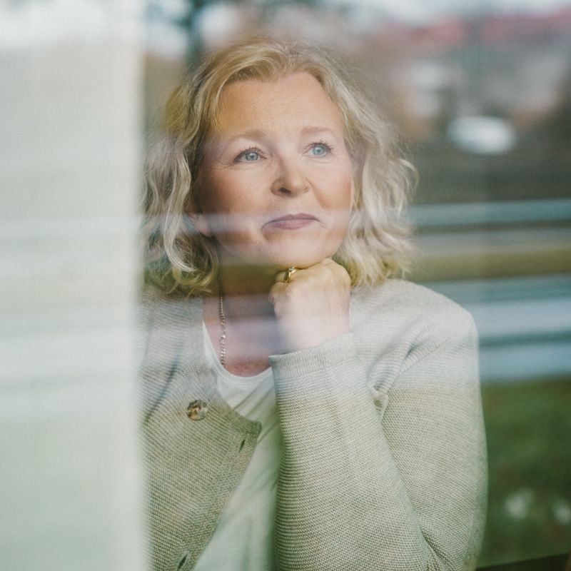 Ett stämningsfullt porträtt av Helene Fritz sett genom en glasruta. Hon vilar hakan i handen och blickar drömskt utåt, vilket skapar en personlig och harmonisk känsla.