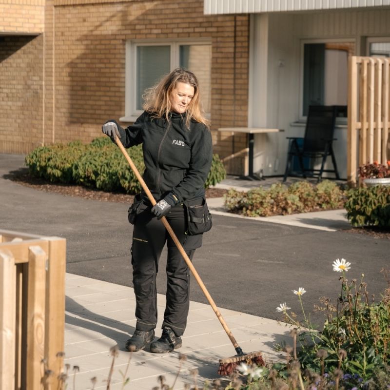 En medarbetare från Fabo sopar en stenlagd gångväg utanför ett bostadshus, vilket visar på det dagliga arbetet med att hålla områdena rena och välskötta.