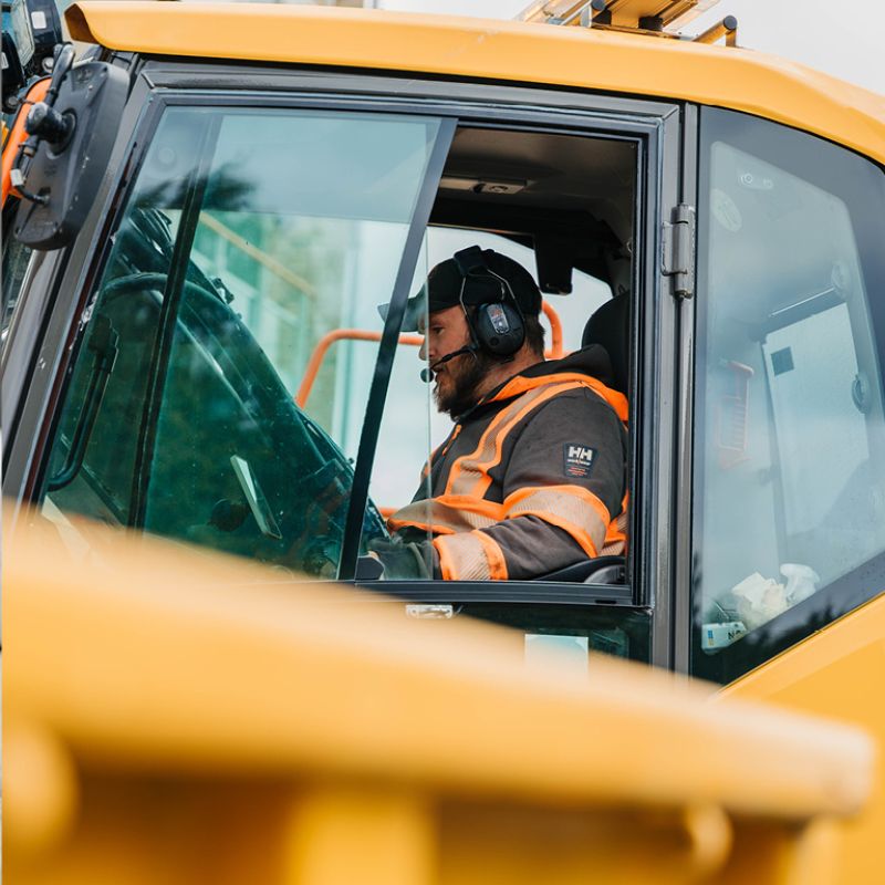 En medarbetare från Markteknik i varselkläder och hörselkåpor sitter koncentrerad i förarhytten på en gul anläggningsmaskin.
