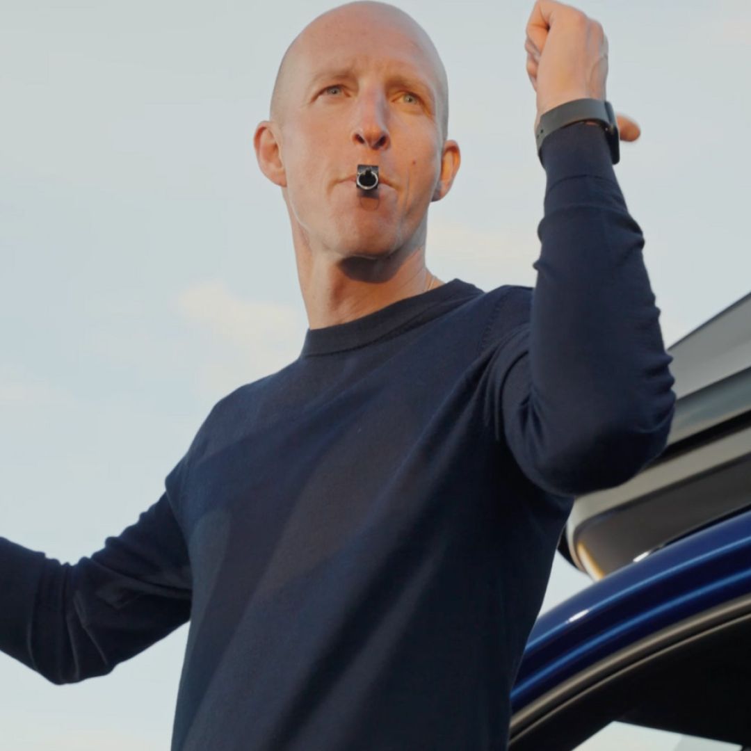 En närbild på en flintskallig domare som blåser i en pipa och sträcker upp armen, vilket indikerar en signal under ett utomhusevenemang som arrangeras av Lunds Bil. Bakom honom syns taket på en blå BMW och en ljus himmel.