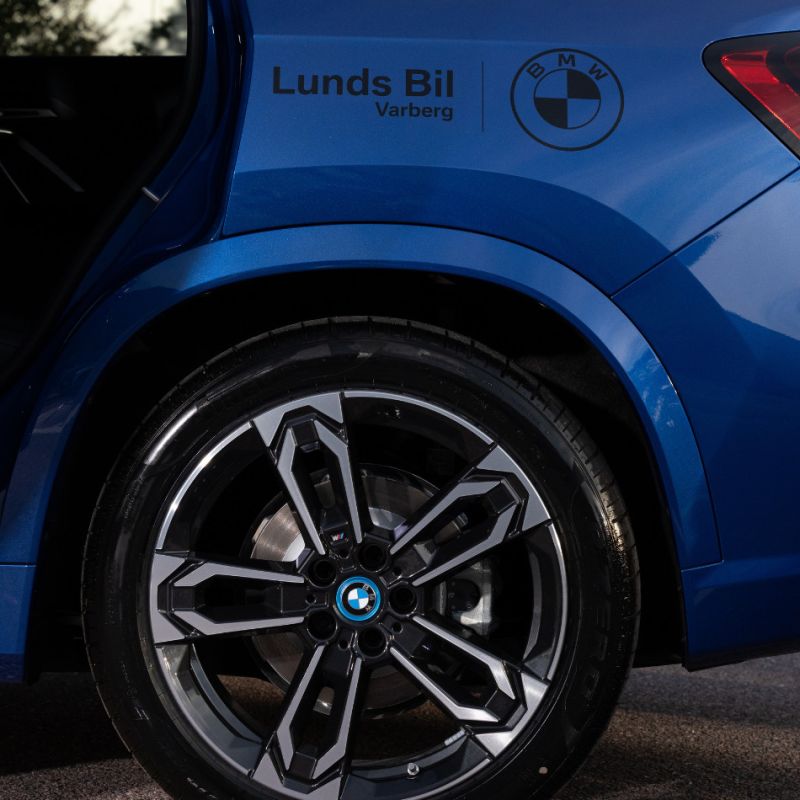 En närbild på bakhjulet och skärmen på en blå BMW. På karossen sitter en svart dekal med texten ”Lunds Bil Varberg” intill BMW-logotypen, vilket visar företagets avsändare på kampanjbilen.