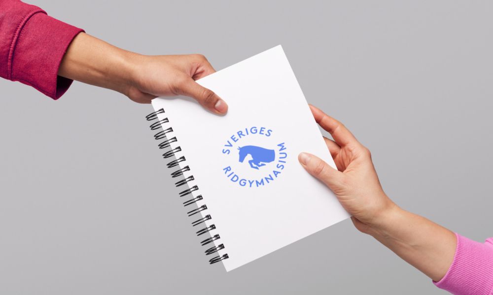En person räcker över ett vitt spiralblock med Sveriges Ridgymnasiums logotyp till en annan person.