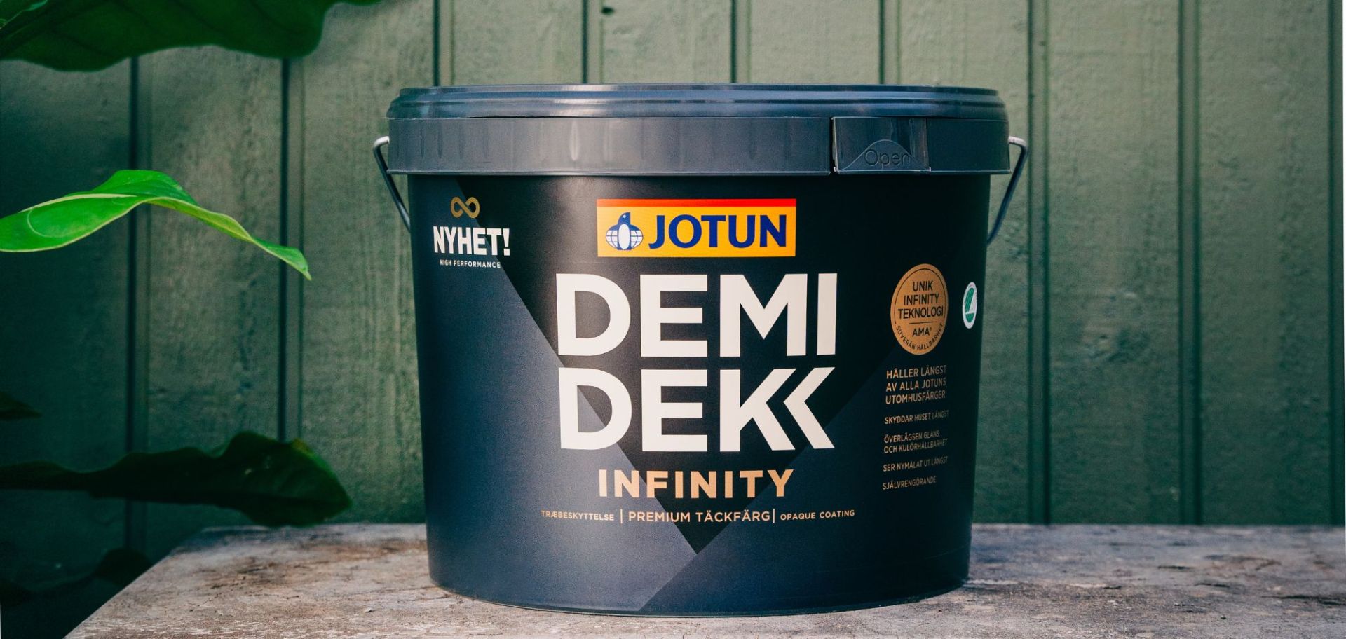  En hink med Jotun Demidekk Infinity täckfärg placerad utomhus framför en grönmålad trävägg.