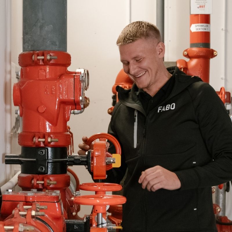  En skrattande medarbetare från Fabo utför underhållsarbete vid ett sprinklersystem med röda rör och ventiler, vilket visar på en positiv arbetsmiljö och teknisk tillsyn.