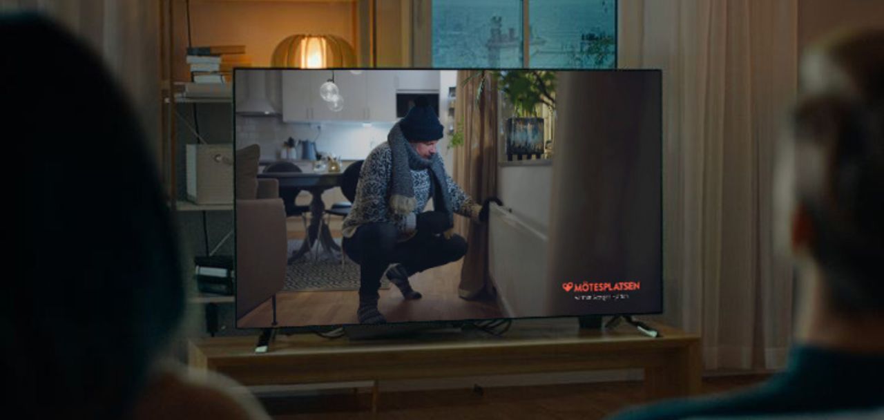  En man i vinterkläder som värmer sig vid ett element visas på en TV-skärm med Mötesplatsens logotyp i hörnet.