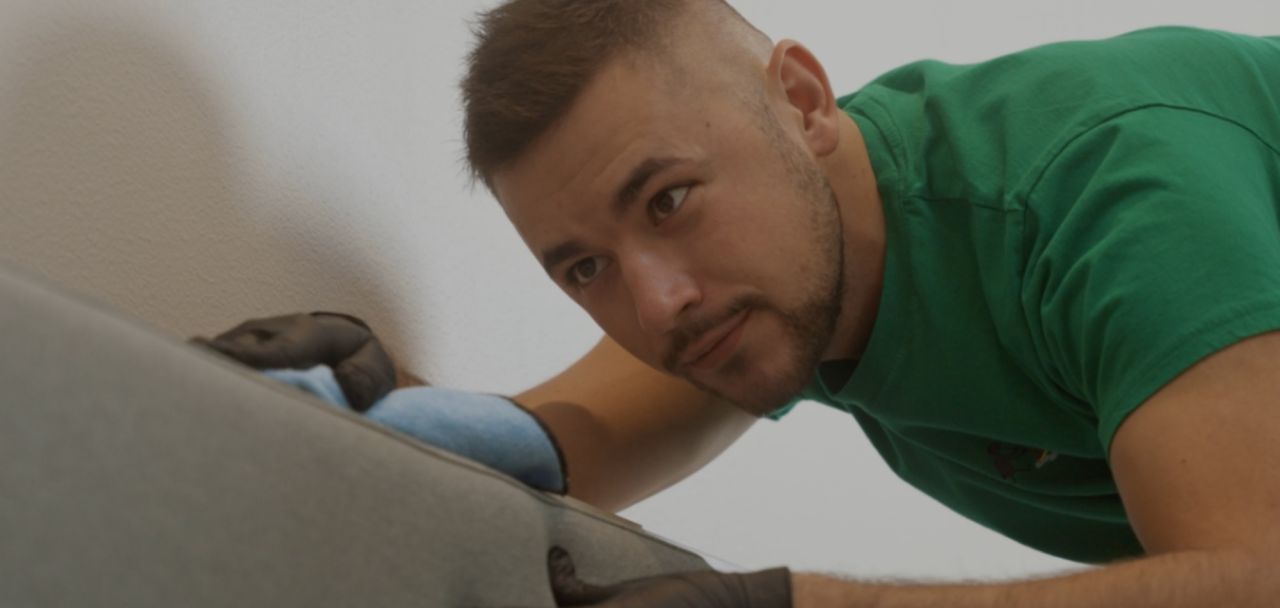 En närbild på en anställd man i grön profil-t-shirt som noggrant rengör en grå möbel. Han bär svarta arbetshandskar och använder en blå putsduk för att torka av ytan med stort fokus.