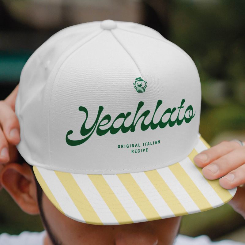 En närbild på en vit keps med Yeahlatos gröna logotyp och texten ”Original Italian Recipe” på framsidan. Skärmen pryds av varumärkets karakteristiska gulvita ränder.