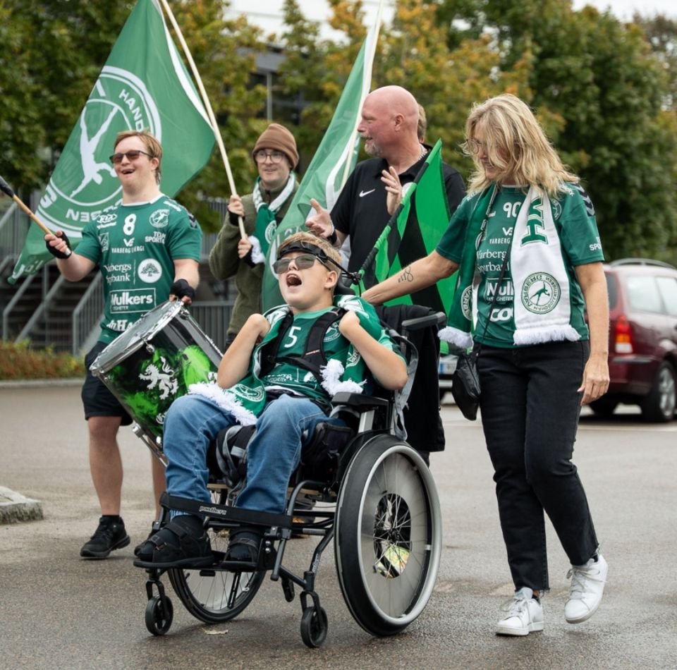  En grupp glada supportrar från HK Varberg, inklusive en pojke i rullstol, hejar och firar med flaggor och trummor.