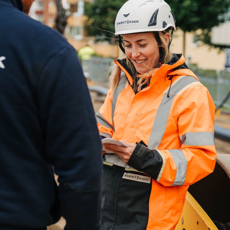 En kvinnlig medarbetare från Markteknik i orange varseljacka och vit skyddshjälm ler under ett samtal med en kollega på en arbetsplats.
