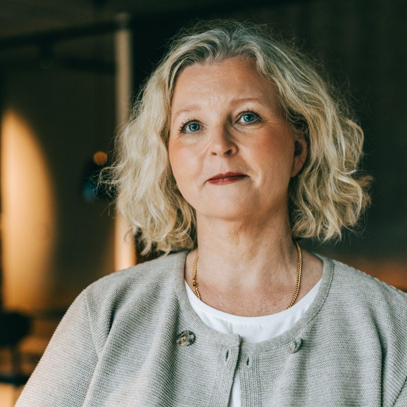 Ett porträtt av Helene Fritz som tittar in i kameran med ett lugnt och professionellt uttryck. Hon har blont, lockigt hår och bär en ljusgrå kofta över en vit topp, med en varm belysning i bakgrunden.