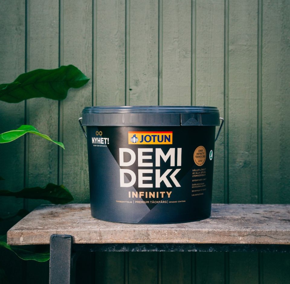 En färgburk från Jotun Demidekk Infinity placerad på en stenbänk framför en grönmålad träfasad.