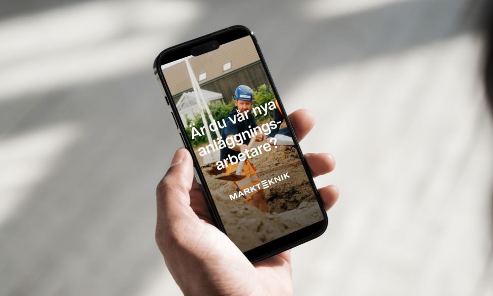 En smartphone visar en jobbannons för Markteknik med texten "Är du vår nya anläggningsarbetare?" och en bild på en roterande grävmaskinshink.
