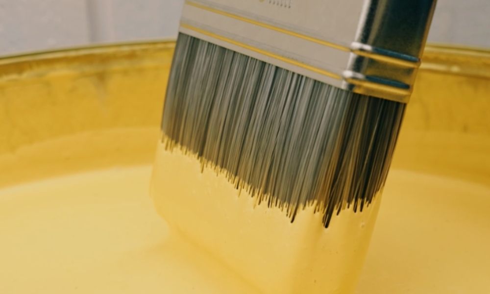Närbild av en målarpensel som doppas i en hink med gul färg.