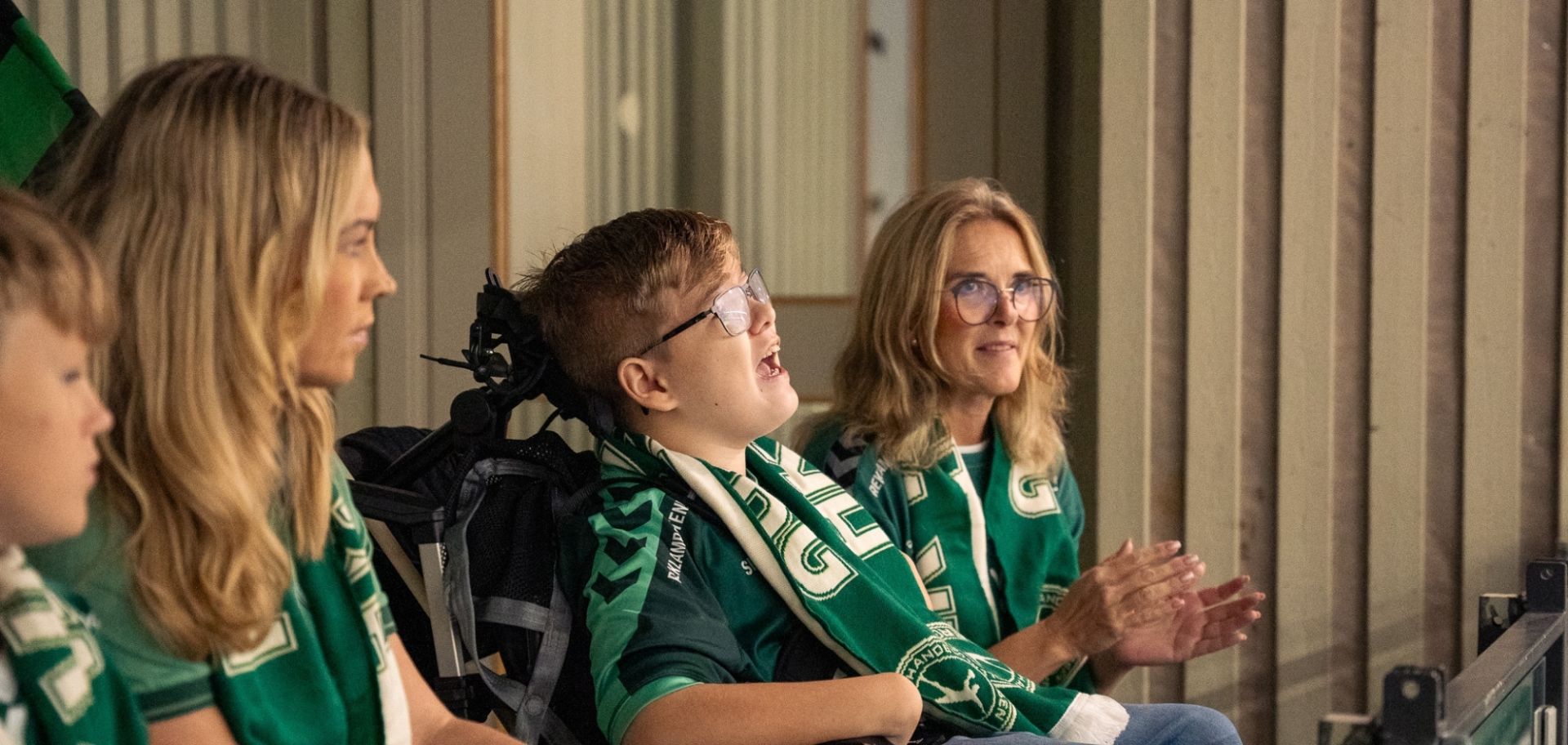  Supportrar i gröna tröjor följer spänningen från läktaren, där en ung kille i glasögon hejar hängivet tillsammans med sin familj.