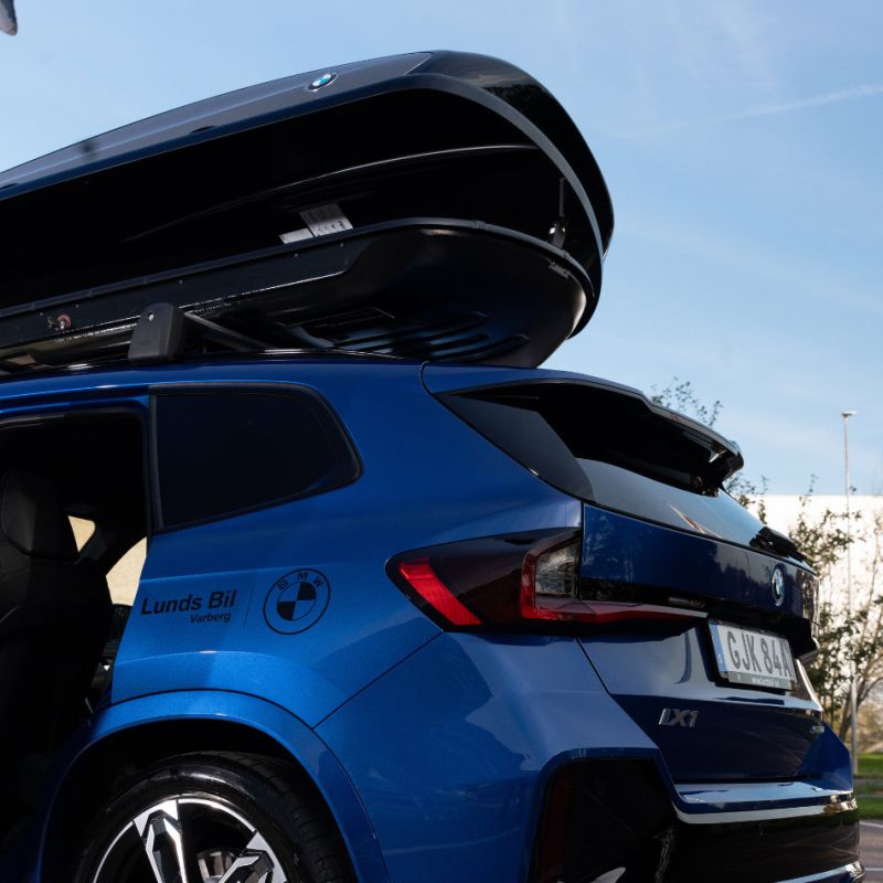 En närbild bakifrån på en blå BMW iX1 med en öppen takbox monterad på takräcket. Bilens sida är profilerad med logotyper för BMW och Lunds Bil Varberg mot en bakgrund av blå himmel och trädtoppar.