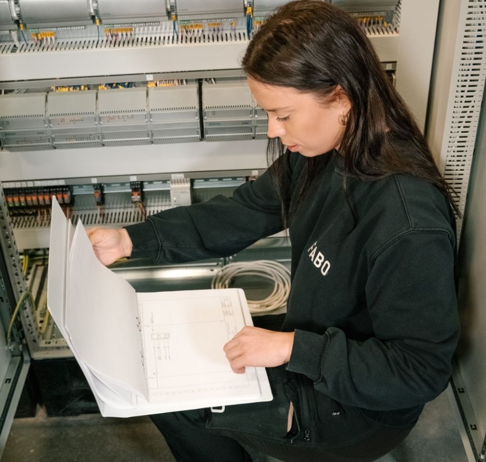  En medarbetare från Fabo går igenom teknisk dokumentation i en pärm framför ett elskåp, vilket visar på precision och tekniskt underhåll i verksamheten.