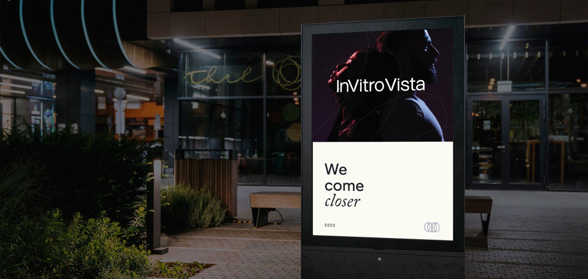 En digital reklamskylt utomhus i kvällsmiljö som visar InVitro Vistas visuella identitet. Affischen är uppdelad i en mörk övre del med en konstnärlig profilbild och logotypen, samt en ljus nedre del med budskapet ”We come closer” och företagets cirkelformade symbol.