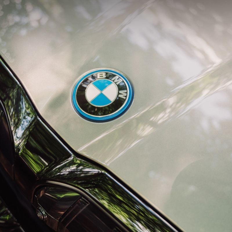  En närbild på BMW-logotypen på motorhuven av en silverfärgad bil, där den blanka lacken reflekterar omgivande grönska.