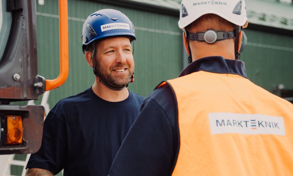  En medarbetare från Markteknik i blå skyddshjälm ler mot sin kollega under ett samtal på arbetsplatsen.